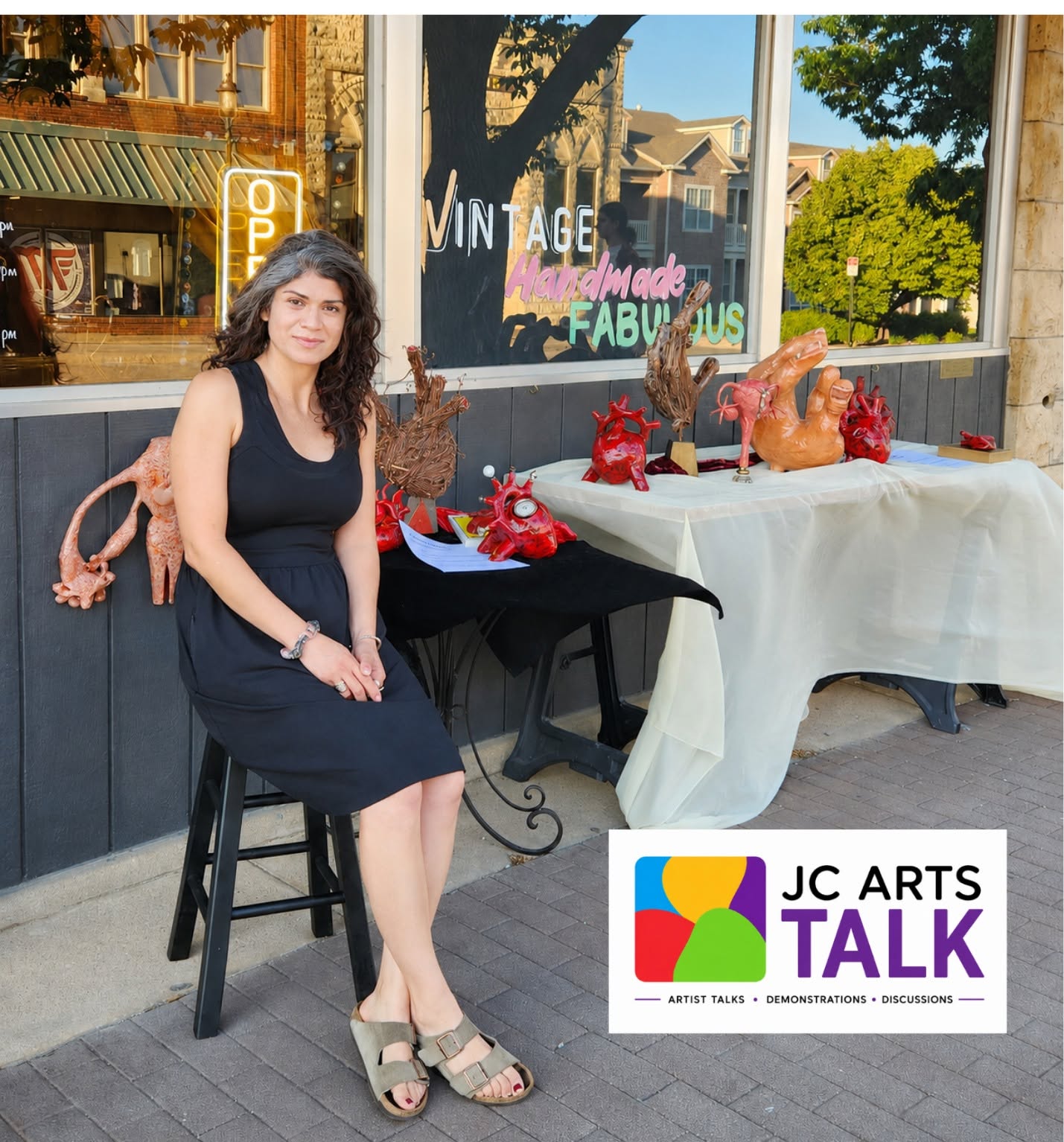 photo of a woman with dark hair wearing a black sundress and sitting on a stool outside of a storefront.