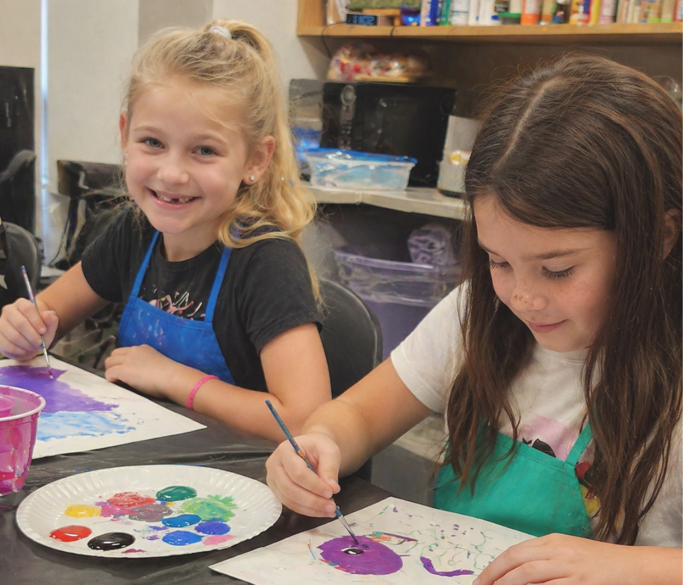 2 female children wearing art aprons painting on white art paper at a table.