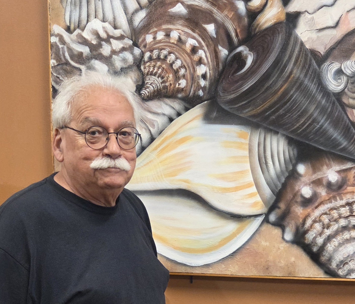 older white man with white hair, white mustache, eye glasses and dark shirt standing beside a painting on a brown wall